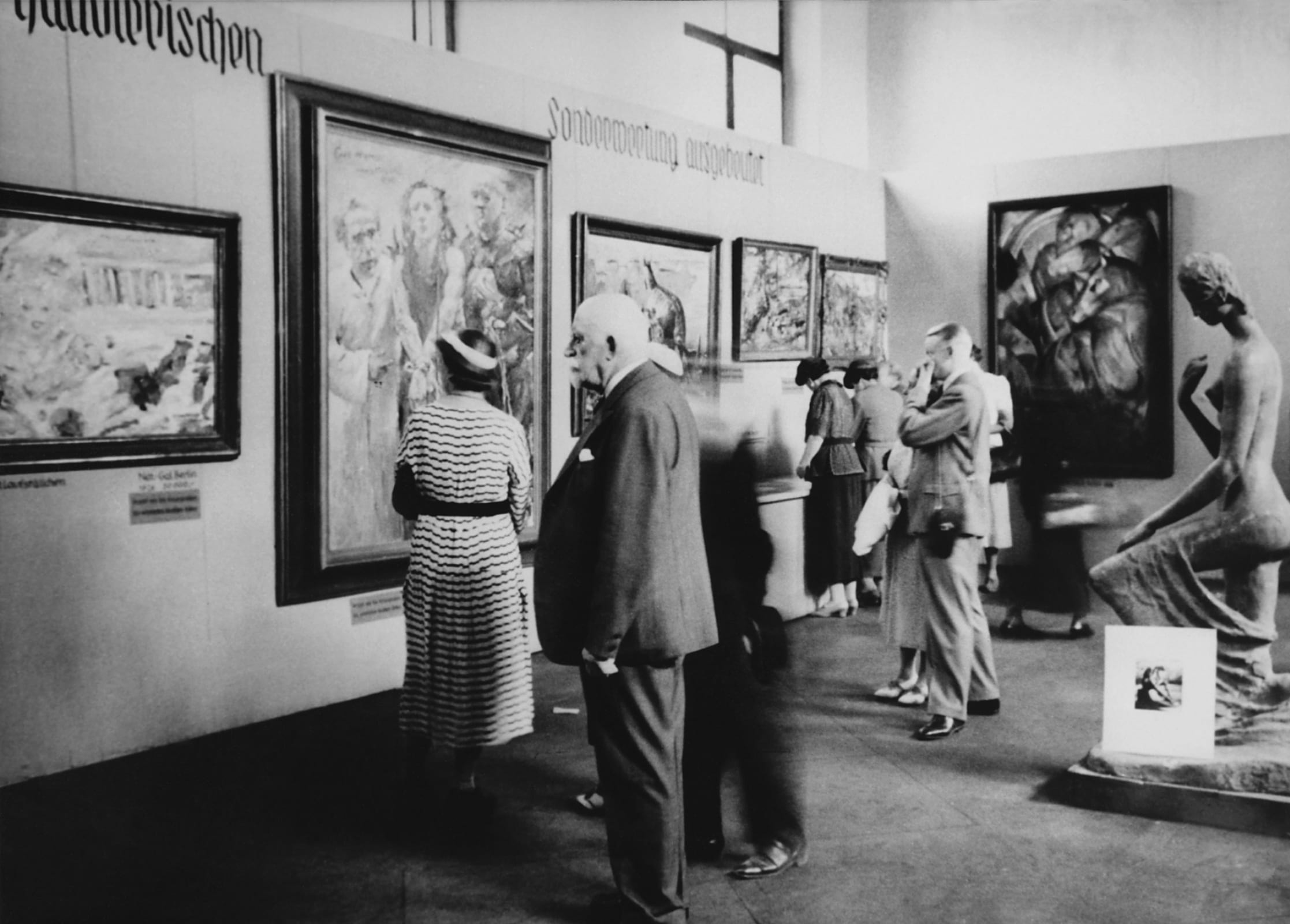Ziyaretçiler, Dejenere Sanat sergisinde. Resimde Lovis Corinth'in Ecce homo'su (soldan ikinci) ve Franz Marc'ın Mavi Atlar Kulesi (sağdaki duvar) ile Wilhelm Lehmbruck'un Diz Çökmüş Kadın heykeli görülüyor. Fotoğraf: MoMA